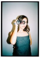 A polaroid of Lea Willms holding a digital video camera. Lea is facing straight ahead, holding the camera directly in front of her face. The screen of the camera is rotated so we can see it. We can see Lea's eye on the screen, directly in front of where her actual eye is.