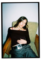 Lea is slouching on a retro armchair. The chair has wooden arms, and olive-green cushions. Lea is wearing all black, and has her hand rested on her stomach as she looks off to the side.
