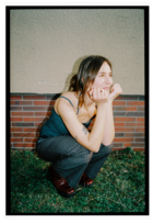 Lea is squatting in a child-like pose on green grass in front of a low red brick wall. She is resting her chin on her hands, gazing at something off to the side.
